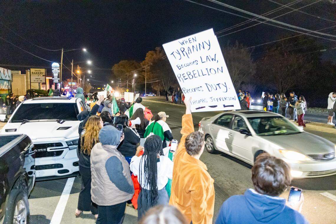 From a slightly elevated perspective, a protester’s back is visible on the right, holding a large white sign above their head. The sign reads in black block letters: “WHEN TYRANNY BECOMES LAW, REBELLION BECOMES DUTY. THOMAS JEFFERSON.” A silver sedan with blurred headlights drives past the protester on a dark street, and other protesters are visible in the background.