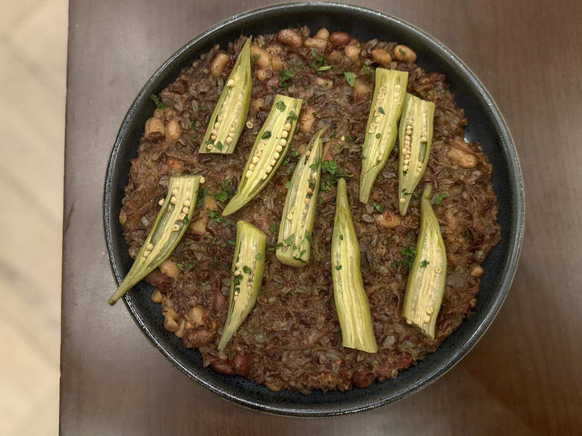 A top-down view of a rustic, dark brown stew served in a shallow dark bowl. The stew appears to contain beans and shredded meat or grains. It is artfully garnished with several sliced, bright green okra pods laid across the top and a sprinkle of fresh chopped herbs.