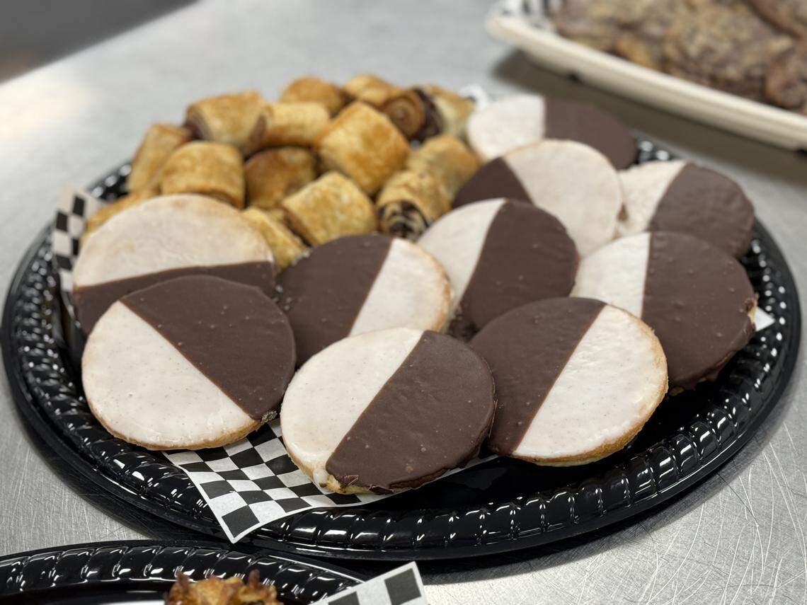 A close-up of a black serving platter filled with Jewish deli desserts. In the foreground are several large black-and-white cookies with their signature half-chocolate, half-vanilla icing. In the background are several small, golden-brown rugelach pastries.
