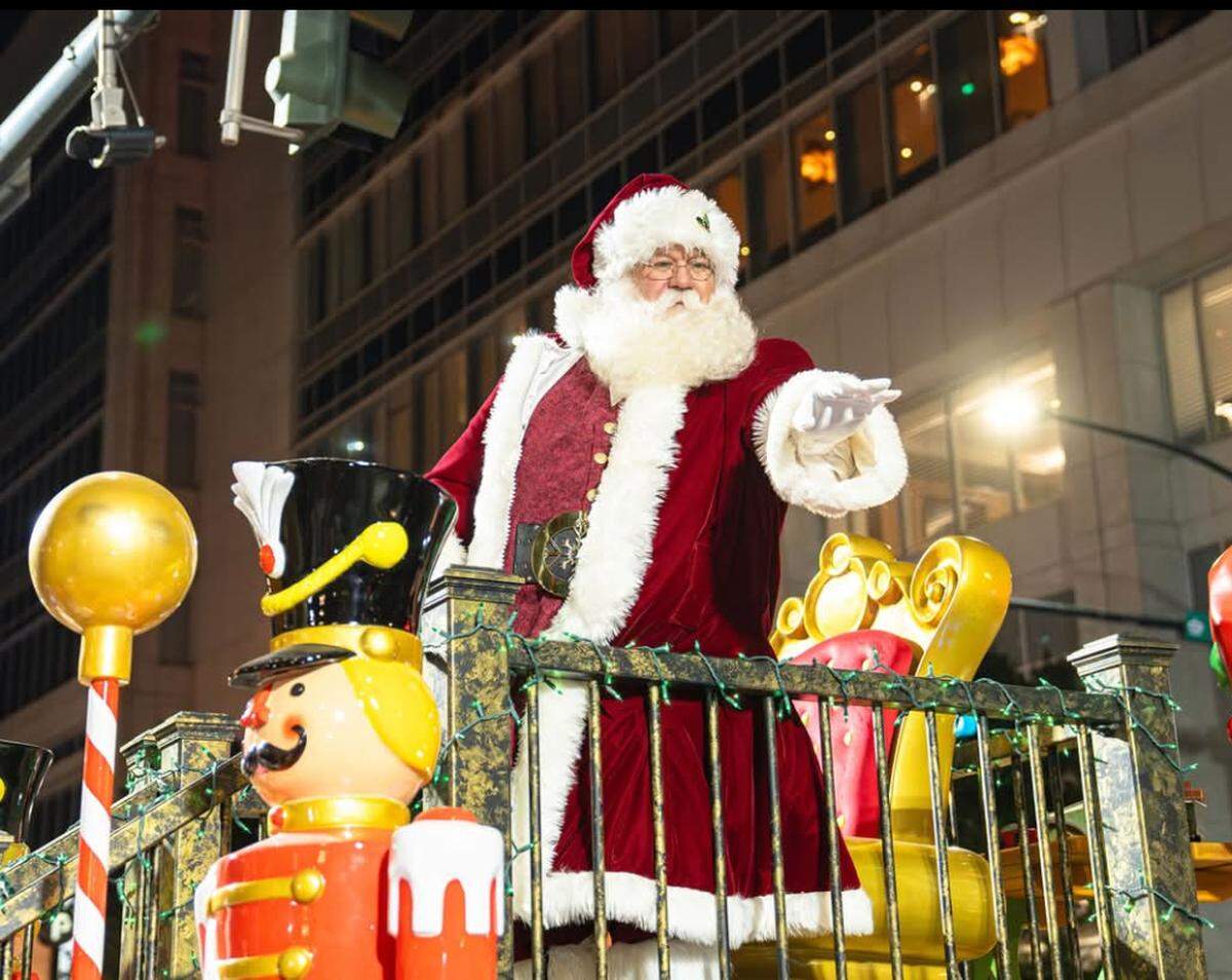 A low-angle shot of a Santa performer in a rich red velvet suit with white fur trim, standing on a decorated parade float. He is gesturing toward a crowd at night, positioned next to a large nutcracker figure and a golden ornate chair.