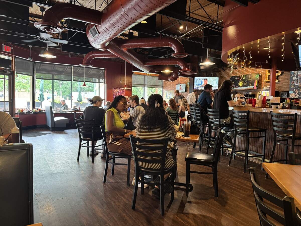 The bustling interior of a casual restaurant and bar with a warm, lively atmosphere. Prominent red exposed ductwork runs across the ceiling. Customers sit at wooden tables in the center and line the wooden bar counter on the right under string lights. Large windows line the left wall, letting in daylight.