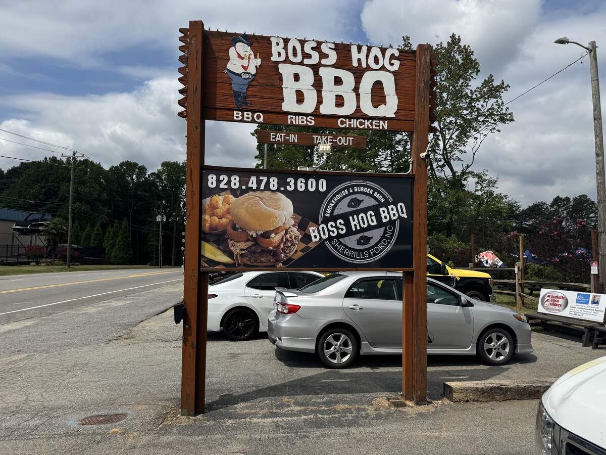 A large roadside wooden sign for “Boss Hog BBQ” in Sherrills Ford, North Carolina. The top portion is made of dark rustic wood with a cartoon pig wearing a cowboy hat and the words “Boss Hog BBQ, BBQ - Ribs - Chicken, Eat-In, Take-Out” in white text. Below it, a black banner shows a photo of a large burger with onion rings next to the restaurant’s circular logo and phone number, 828.478.3600. Two silver cars are parked directly behind the sign.
