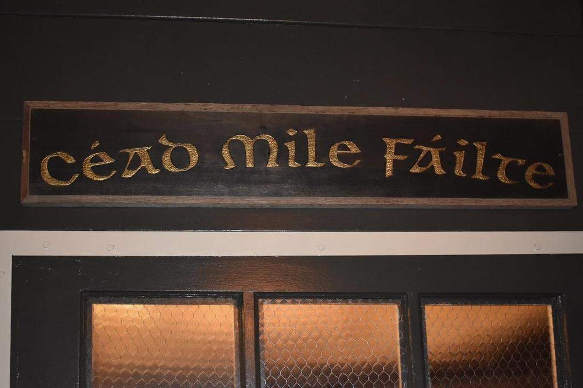 A close-up, straight-on shot of a black wooden sign with a light wood frame. The sign displays the Irish phrase “Céad Míle Fáilte” in ornate gold Celtic lettering, mounted above a dark-painted door with small, square, textured windows.