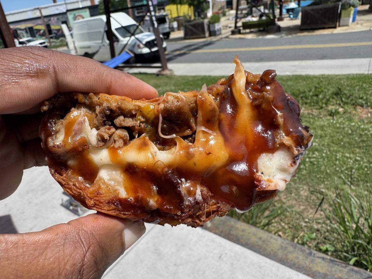 A close-up, cross-section view of a savory eggroll being held up. The crispy golden-brown shell is filled with melted white cheese and seasoned ground meat, all topped with a thick, glossy dark barbecue sauce. The background shows a sunlit outdoor area with a street and sidewalk.