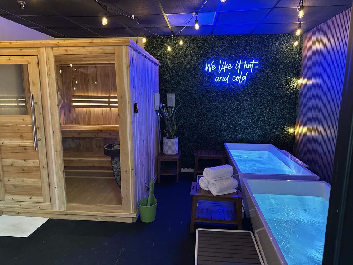 A recovery room with a large wooden sauna on the left and two white cold plunge tubs on the right. A blue neon sign reading “We like it hot ... and cold” is mounted on a dark green textured wall, with string lights hanging from the ceiling.