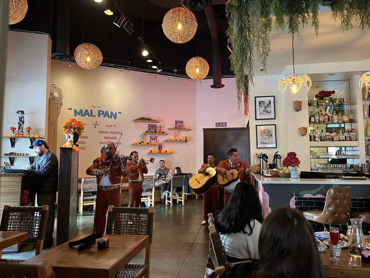 A live mariachi band in traditional brown uniforms performing inside a brightly lit restaurant named Mal Pan, featuring woven spherical pendant lights, a bar area, and dining patrons.