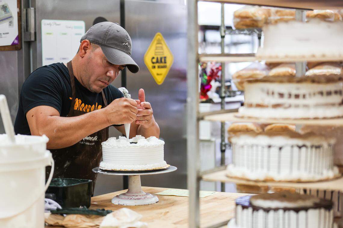 A baker wearing a gray baseball cap and a dark apron carefully piping white frosting into a round, multi-layered white cake on a stand in a professional kitchen or bakery. A tiered metal shelf holding several other partially finished cakes is visible on the right.