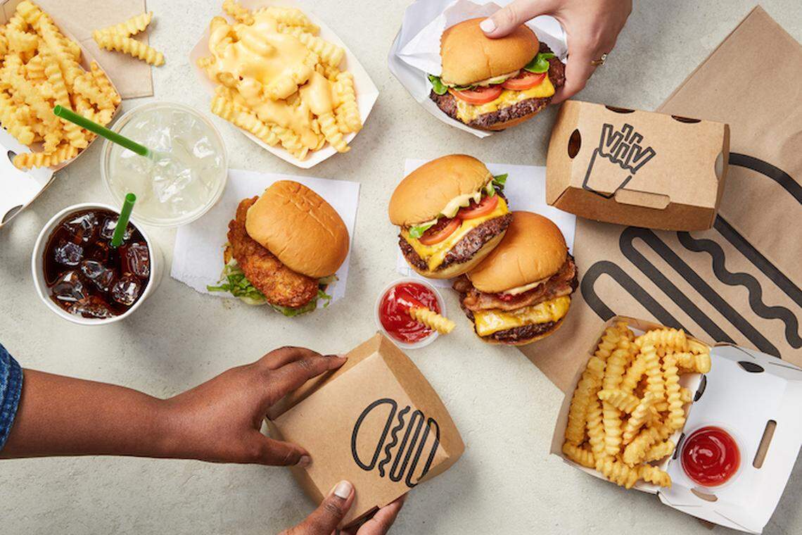 An overhead view of a large delivery spread featuring multiple burgers, crinkle-cut fries with cheese sauce, and drinks arranged on a light-colored tabletop. Hands are visible reaching into the frame to grab a burger and move a branded cardboard delivery box.