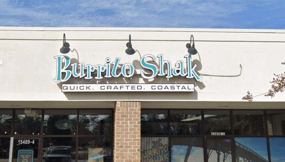 The exterior of a “Burrito Shak” restaurant displays a large, stylized teal and white sign mounted on a cream-colored wall. Below the brand name, a smaller black bar features the words “QUICK. CRAFTED. COASTAL.” in clean white capital letters. Three black gooseneck lamps hang above the sign, illuminating the storefront located in a modern shopping center.