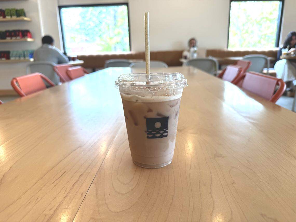 A clear plastic cup of iced latte with a beige paper straw, centered on a light wooden table. The cup features a small, black and white circular sticker logo of waves under a sun or moon. The background is slightly blurred, showing the interior of the coffee shop with people seated at tables, a long, upholstered bench seat, and two large square windows with views of greenery outside.