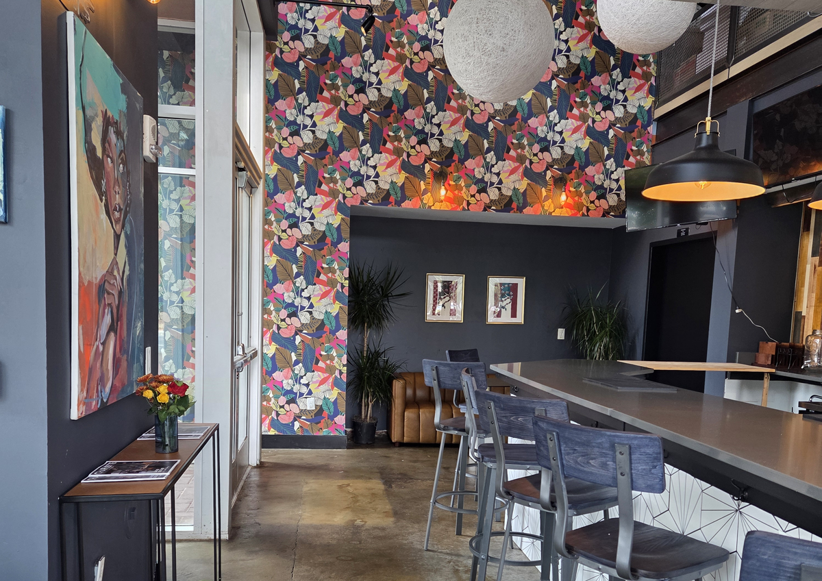 This image captures a view down the length of the cafe’s bar area, featuring dark gray walls and a bold, colorful floral-patterned accent wall extending up to the high ceiling. In the foreground, there are dark wooden bar stools pulled up to the counter, with a glimpse of the green velvet table seating on the left.