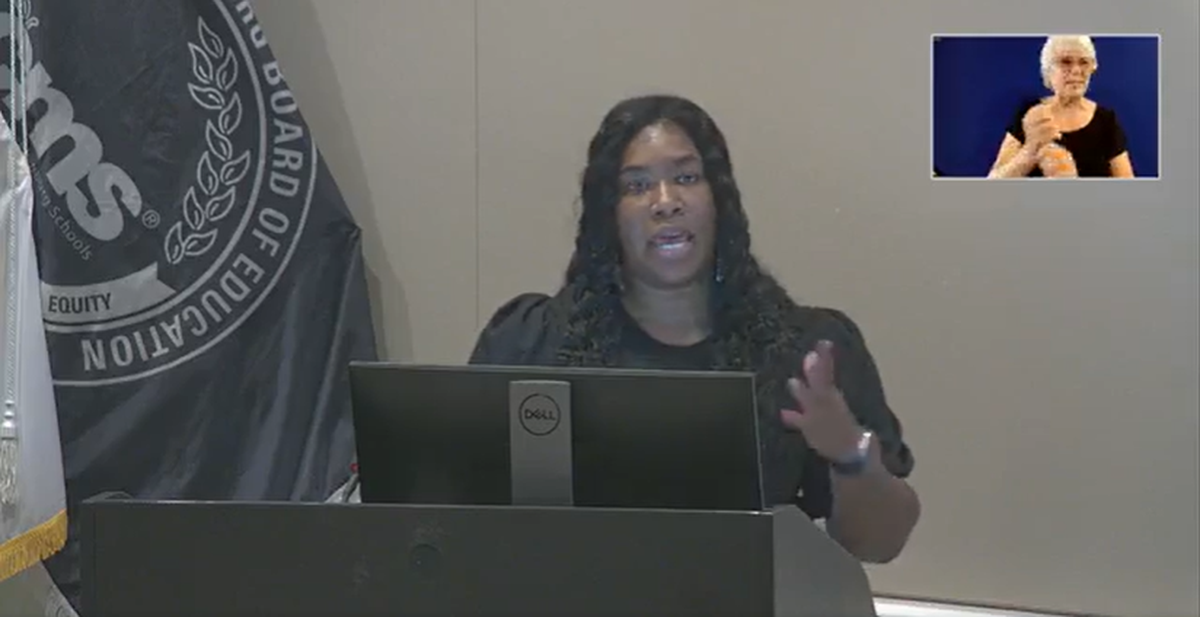 A Black woman speaks from a podium in front of a gray background.