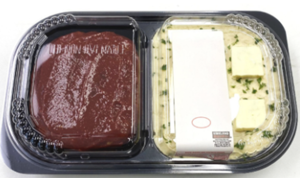 A top-down shot shows a Kirkland Signature pre-packaged meal in a black plastic container with two clear-lidded compartments. The left side contains a meatloaf topped with a thick red glaze, while the right side features mashed potatoes garnished with fresh herbs and two pats of butter.