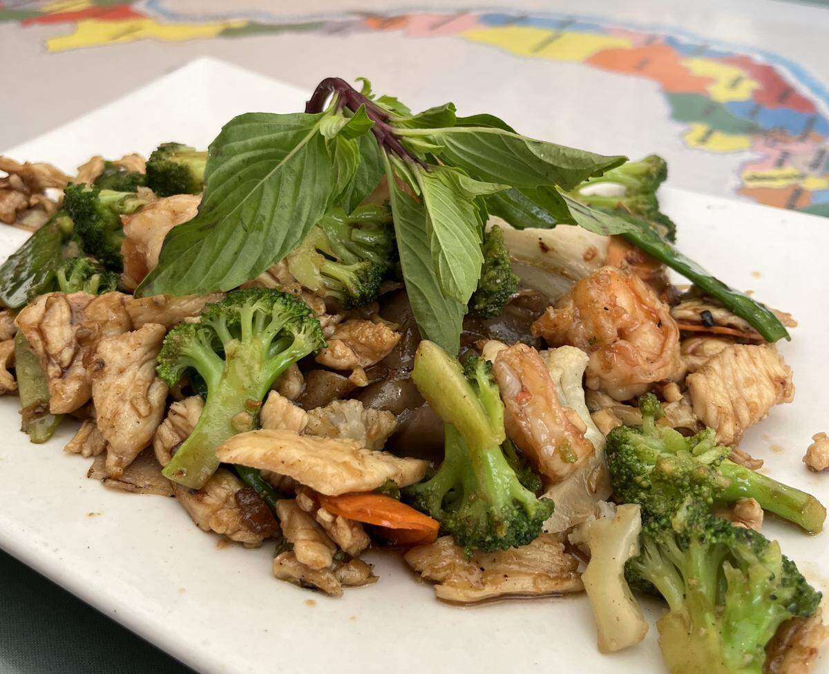 A close-up, angled shot of a white rectangular plate filled with a stir-fry dish, placed on a colorful map-themed tablecloth. The dish contains pieces of chicken, shrimp, broccoli florets and other vegetables, and wide rice noodles, all coated in a sauce and garnished with a large, fresh sprig of Thai basil.