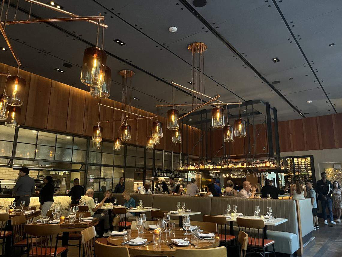 A panoramic shot of a large, modern, and bustling restaurant dining room. The room has a high ceiling with many unique, glass and metal pendant light fixtures hanging over the tables and bar area. The decor features wood paneling on the walls, and a variety of seating is visible, including long booths and round and square tables with wooden chairs. Patrons are seated throughout the restaurant, and an open kitchen area is visible to the left.