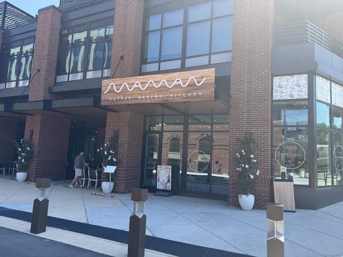 The exterior facade of a modern cafe named “maman COFFEE . BAKERY . KITCHEN,” located in a multi-story red brick building. The storefront features a prominent wooden sign with the cafe’s cursive white logo and a large glass entryway displaying the words “COFFEE,” “BAKERY,” and “KITCHEN” on the panes. The clean concrete sidewalk out front is flanked by dark metal bollards and decorated with potted topiary trees bearing white flowers. A patron is visible walking toward an outdoor seating area tucked under the building’s overhang to the left.