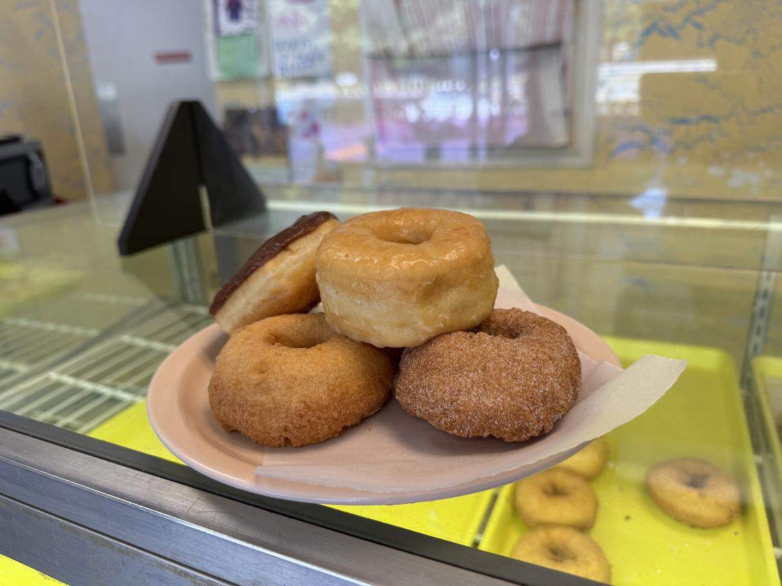 Inside a donut shop’s glass display case, a small plate is presented with a sample of five donuts. A shiny glazed donut sits atop two plain cake donuts and a cinnamon-sugar coated donut. A chocolate bar donut leans against the stack in the back. Below the plate, a yellow tray with more donuts is visible.