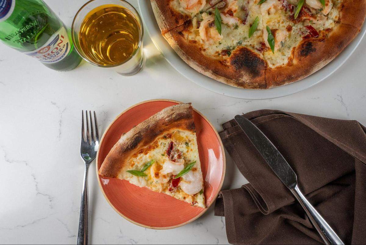 An overhead shot of a slice of shrimp pizza on an orange plate, with the rest of the pizza, a glass of beer, and a Peroni bottle on a white marble table.