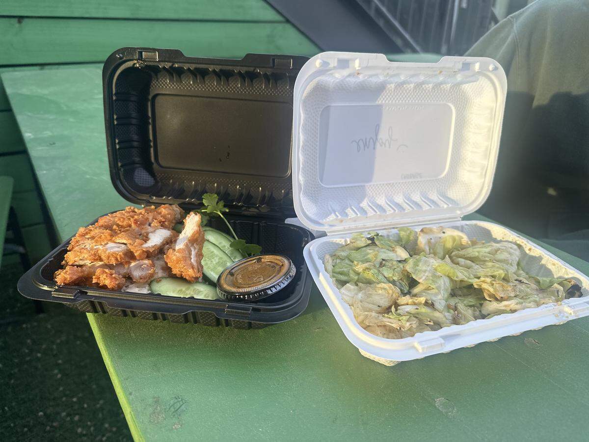 Two open takeout containers sit on a bright green outdoor table, showcasing a hearty meal. The black container on the left holds Khao Man Gai Tod—crispy fried chicken sliced over a bed of seasoned rice, garnished with fresh cucumber slices, cilantro, and a small cup of savory dipping sauce. The white container on the right is filled with sautéed cabbage, lightly charred and seasoned. The inside of the white lid features a friendly, handwritten “Enjoy :)” message.