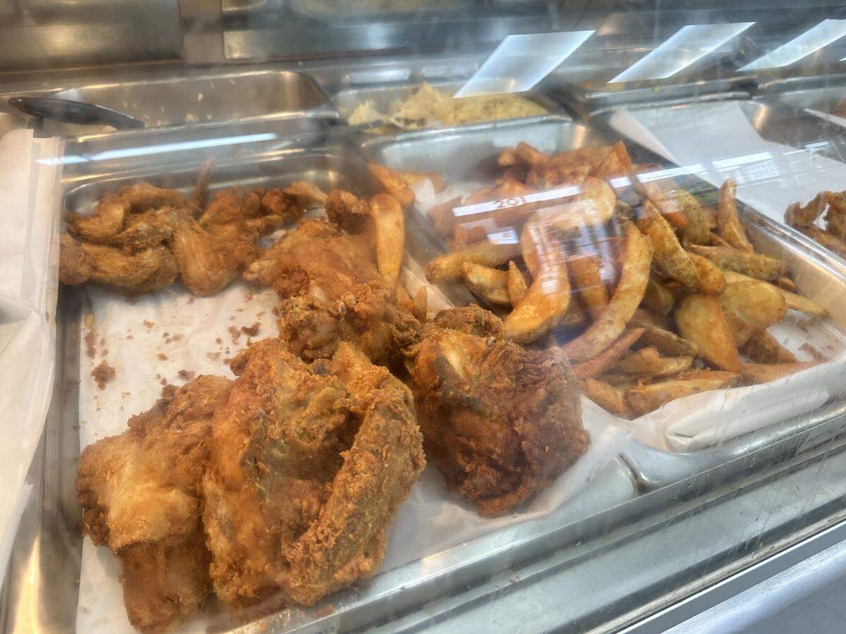 A close-up, angled shot looking down into a glass-and-metal hot food display case. The display features two large, paper-lined metal trays: one in the foreground filled with large, golden-brown pieces of fried chicken, and one to the right filled with a large pile of seasoned potato wedges.