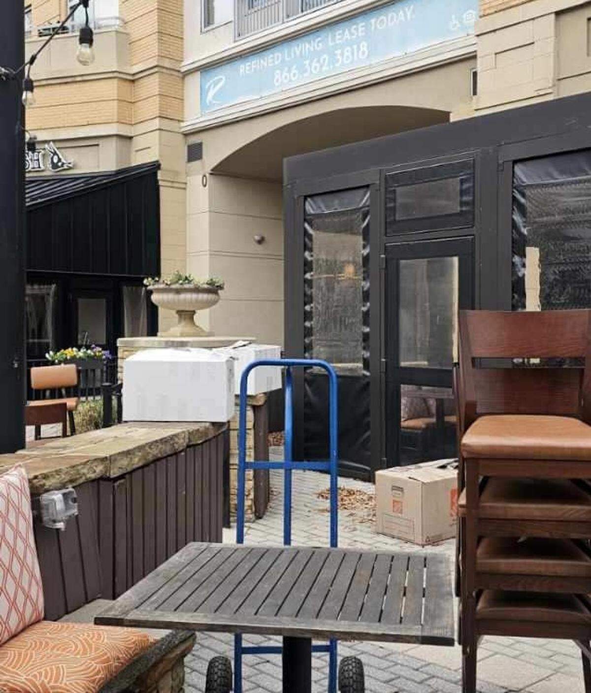 A slightly high-angle shot of a brick paved outdoor patio with stacks of chairs, boxes, and a blue hand truck dolly in the foreground. In the background, there is a closed black structure and an archway entrance to a larger commercial building.