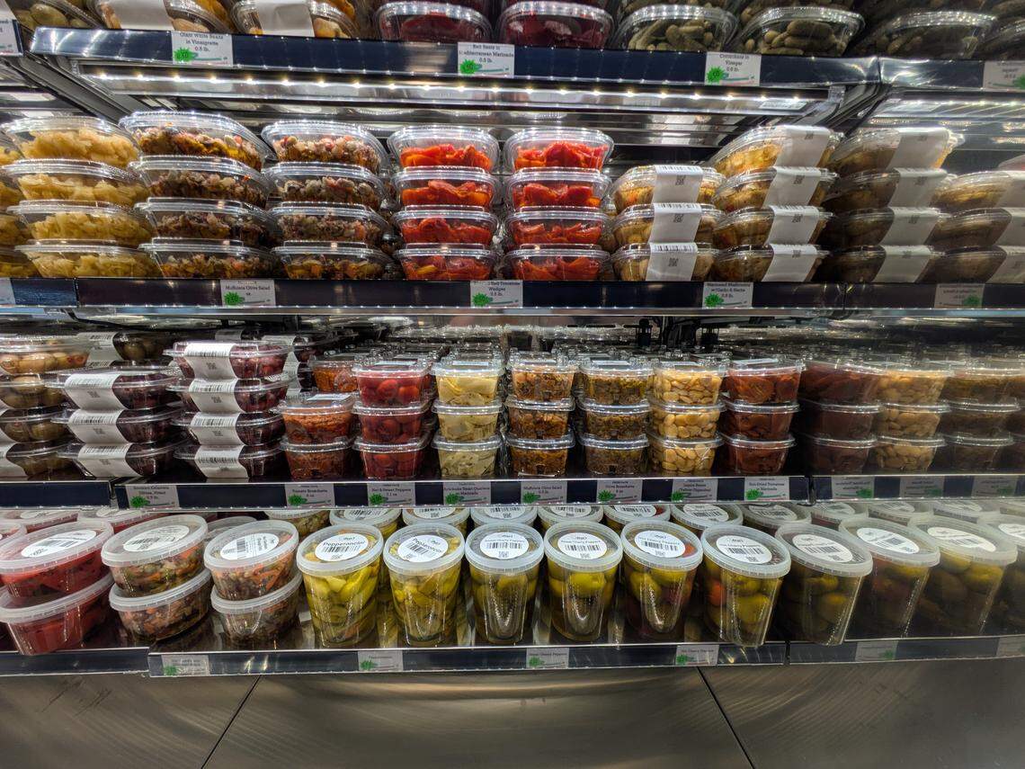 A refrigerated display shelf stocked with various pre-packaged Italian appetizers and deli items. Neatly stacked clear plastic containers hold olives, marinated peppers, mushrooms, artichoke hearts, and sun-dried tomatoes.