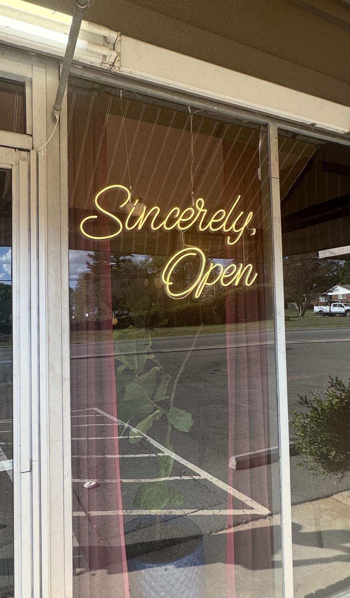 A close-up of a glass storefront door featuring a glowing yellow neon sign that says “Sincerely, Open” in a cursive font. Through the glass, a parking lot and a green potted plant are visible.
