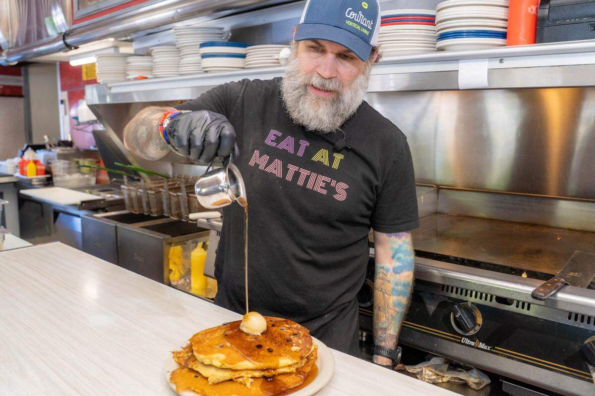 A man with a full gray beard, wearing a baseball cap and a black t-shirt that reads “EAT AT MATTIE’S,” pours syrup from a small metal pitcher onto a stack of pancakes topped with a pat of butter. The scene takes place in a diner kitchen, with stacks of plates and a grill visible in the background.