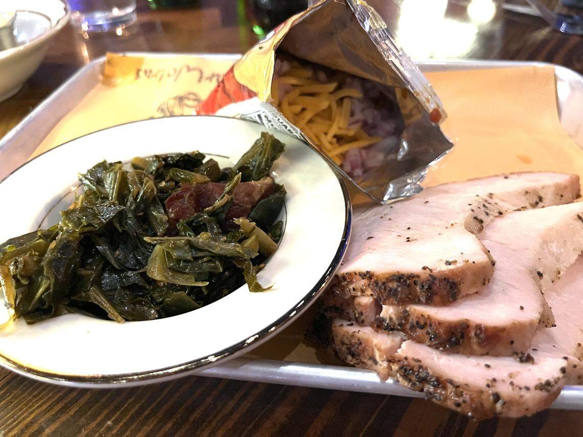 A rustic meal is presented on a paper-lined metal tray on a dark wood table. The platter includes thick slices of smoked turkey, a separate small plate of braised collard greens, and an open bag of Fritos that has been filled with toppings to create a Frito pie.