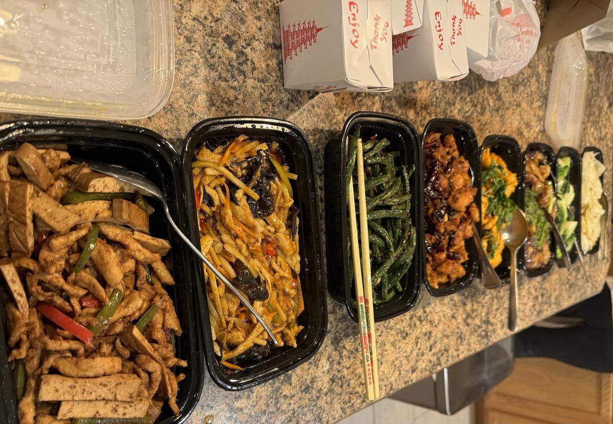A generous takeout meal from Lee Cafe is spread across a countertop. In the foreground, a row of open black containers showcases dishes such as stir-fried tofu and peppers, a spicy shredded pork dish, green beans, and a saucy fried chicken dish. Classic white paper takeout boxes are stacked in the background.