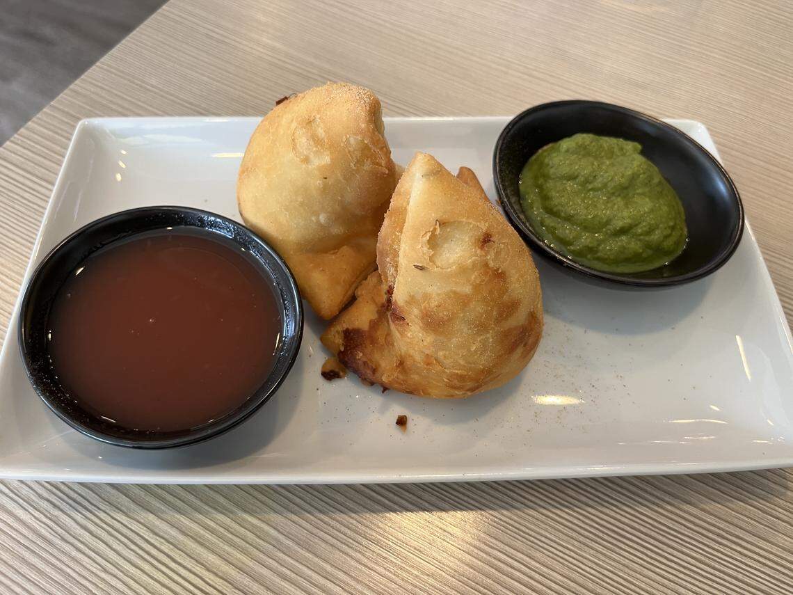 A close-up of two triangular, golden-brown samosas served on a rectangular white plate. The samosas are flanked by two small, black bowls of dipping sauces: a reddish-brown tamarind chutney on the left and a bright green cilantro/mint chutney on the right. The plate is on a light-colored wooden table.