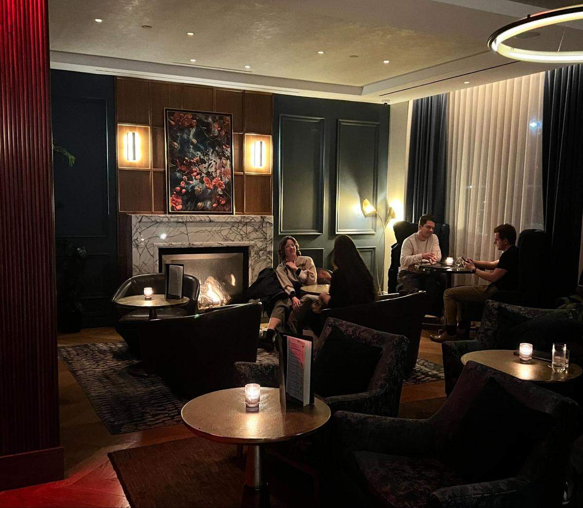 A low-lit, sophisticated lounge area with a cozy fireplace. The room has dark walls with wood paneling and a large white marble fireplace with a roaring fire. A dark, framed floral painting hangs above the mantle, flanked by two vertical light fixtures. Guests are seated in plush dark armchairs and sofas around small round tables with candles. On the left, a couple is seated by the fire, while two men are seated further back, engaged in conversation. Large windows are covered by dark blue drapes and sheer white curtains. The floor is covered with a dark patterned rug. The overall atmosphere is intimate and elegant.