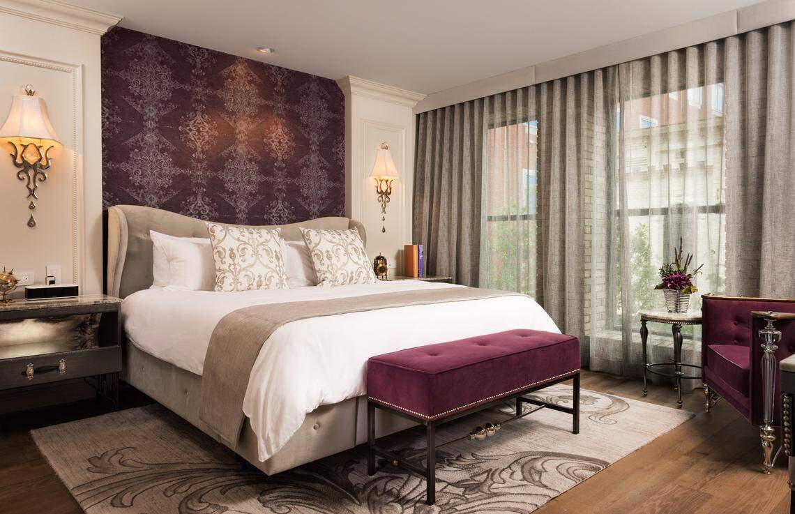 A luxurious hotel bedroom featuring a large bed with white linens, framed by deep purple damask wallpaper and cream-colored paneling. A purple velvet bench sits at the foot of the bed, and sheer gray curtains hang over the window. Ornate wall sconces flank the upholstered headboard.