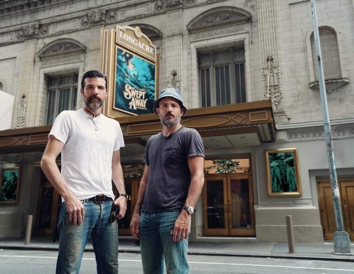Seth Avett, left, and his brother Scott Avett, in New York City in front of the marquee for “Swept Away.” The shipwreck survival musical uses songs from The Avett Brothers catalog.