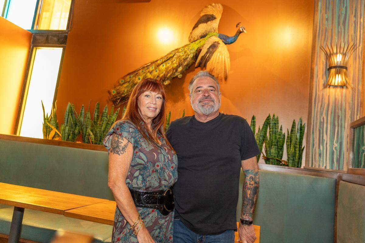 A couple stand together in a restaurant with a distinctive peacock decor. The person on the left has reddish-brown hair, is smiling and wearing a patterned dress with a wide black belt and has visible arm tattoos. The individual on the right, with a grey beard, is wearing a black t-shirt and jeans, and has visible arm tattoos as well. Behind them, a taxidermied peacock with its wings spread is mounted on an orange wall, with its long, vibrant tail feathers extending downwards. A row of snake plants is arranged along a green booth in the background, and a warm-toned light fixture is on the right wall.