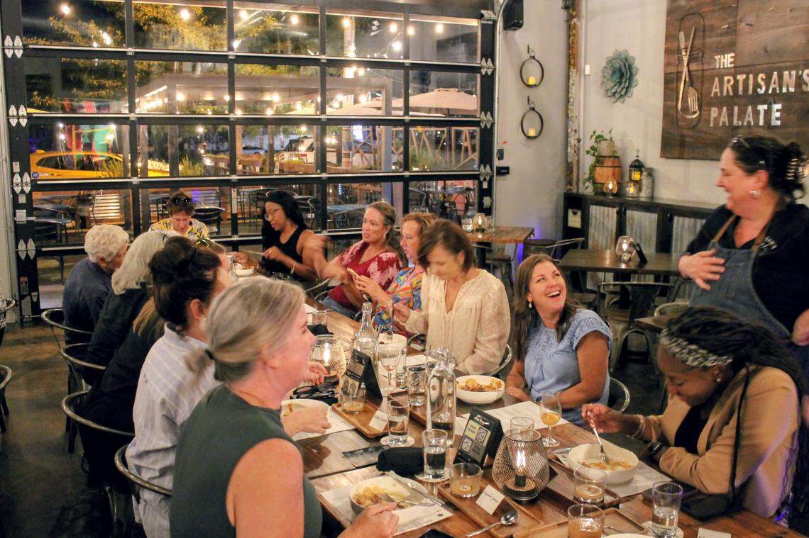 An inviting scene inside a warmly lit, rustic restaurant. A diverse group of women are gathered around a long wooden dining table, sharing a meal and animated conversation. Large garage-door windows in the background show a patio and the street outside at night. A sign on the wall clearly reads “The Artisan’s Palate.”