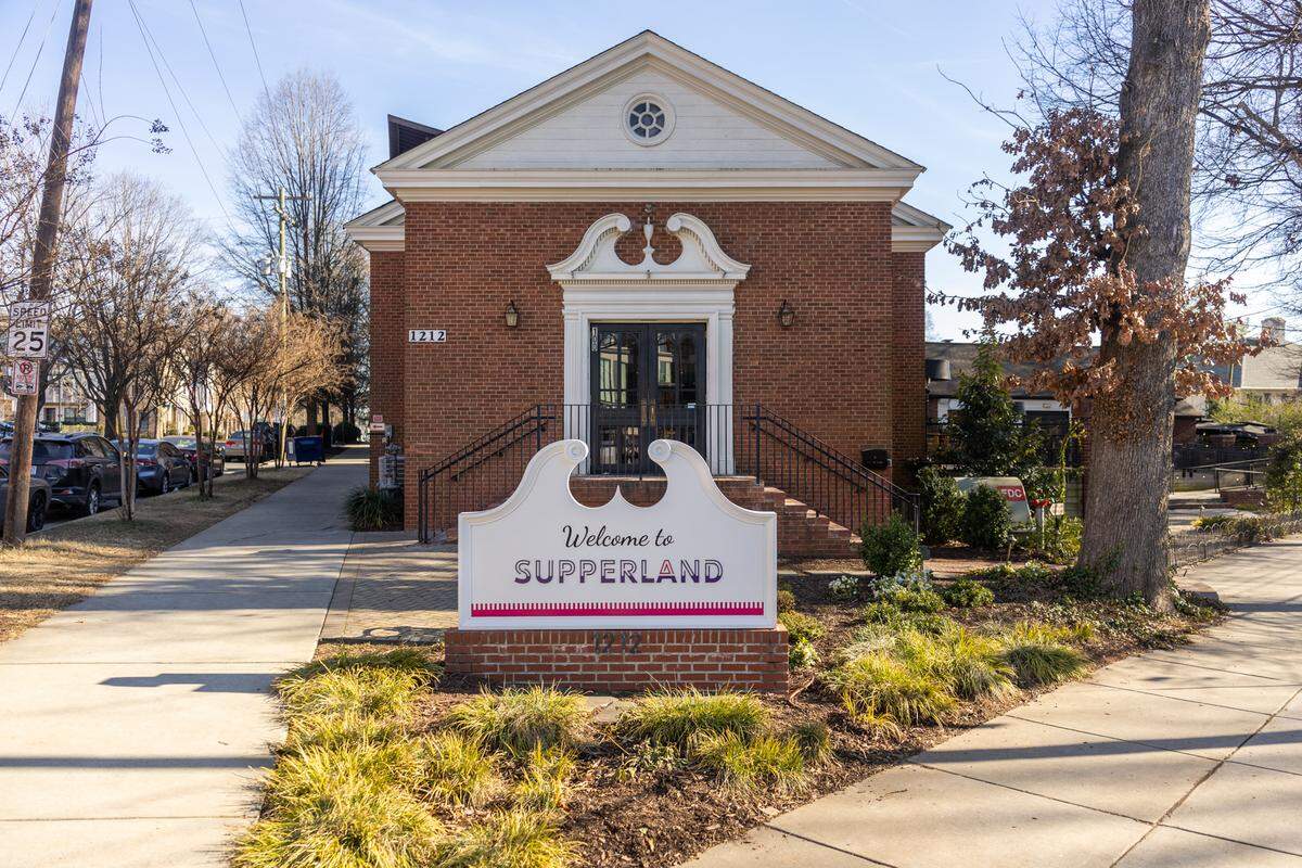 A straight-on, eye-level shot of the “Welcome to SUPPERLAND” sign in the foreground, with the restaurant’s symmetrical, one-story brick building in the background. The building features a gabled roof, white trim, and a central entrance with black double doors, accessed by a set of stairs with black railings. The scene is set on a sunny day with bare-branched trees, a sidewalk, and a street visible on the left.