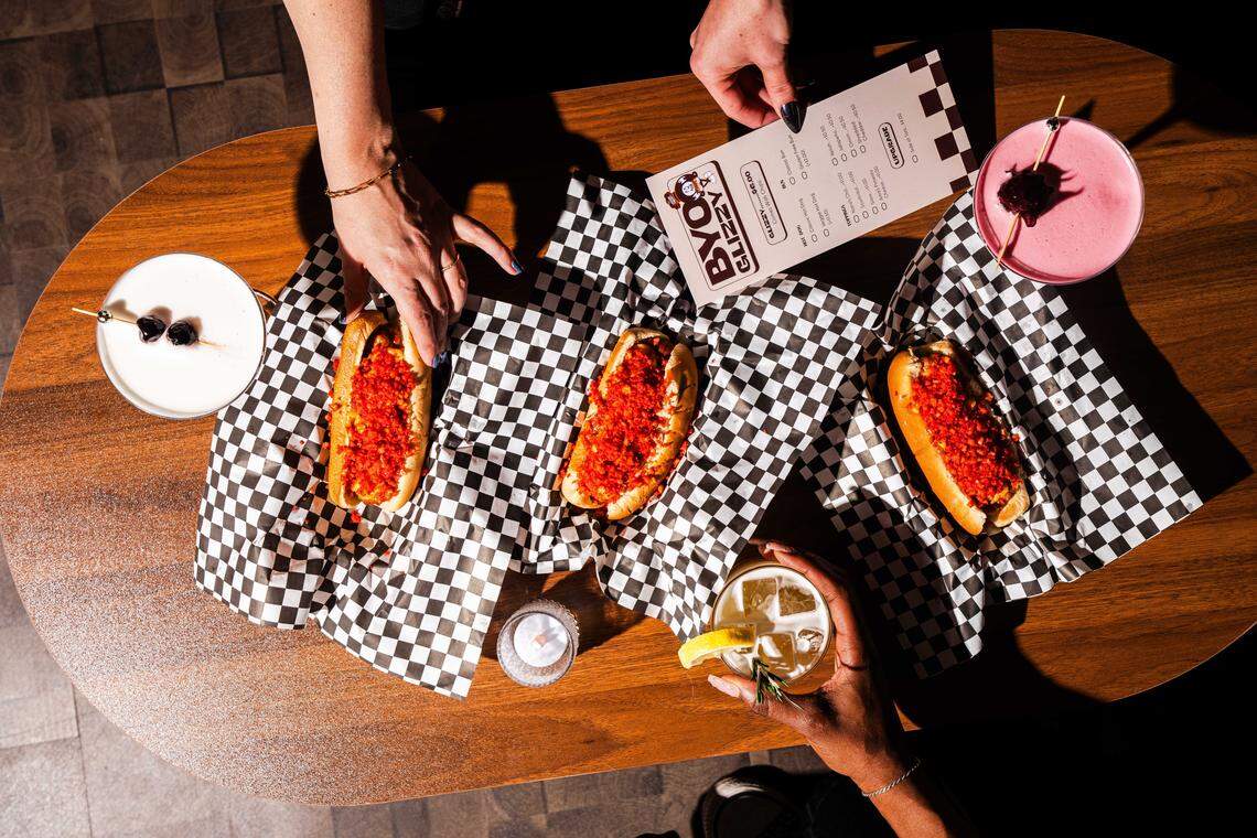 A high-angle, flat-lay shot of a dark wood table featuring three hot dogs topped with bright red, crunchy crumbles, each served on black-and-white checkered deli paper. The spread includes three colorful cocktails—one pink, one white, and one amber—and a small white candle. Several hands are visible in the frame: one reaching for a hot dog, another holding a drink, and a third holding a “BYO Glizzy” menu card.