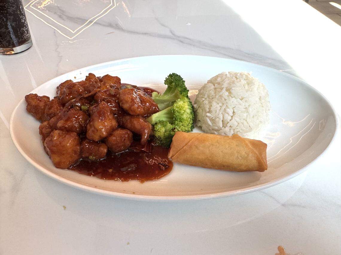 A white oval plate featuring a serving of glossy orange chicken in a dark sauce, a scoop of white rice, two pieces of steamed broccoli, and a single golden spring roll. The plate is set on a white marble tabletop with a dark soda visible in the upper left corner.
