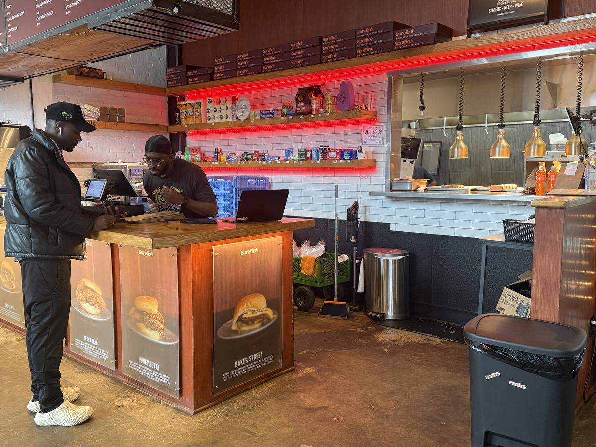 Behind the counter, white subway tiles are illuminated by red neon lighting. Shelves are stocked with convenience items, including household cleaners, paper towels, and snacks like candy and chips. A staff member is visible in the open kitchen area, which features industrial heat lamps.