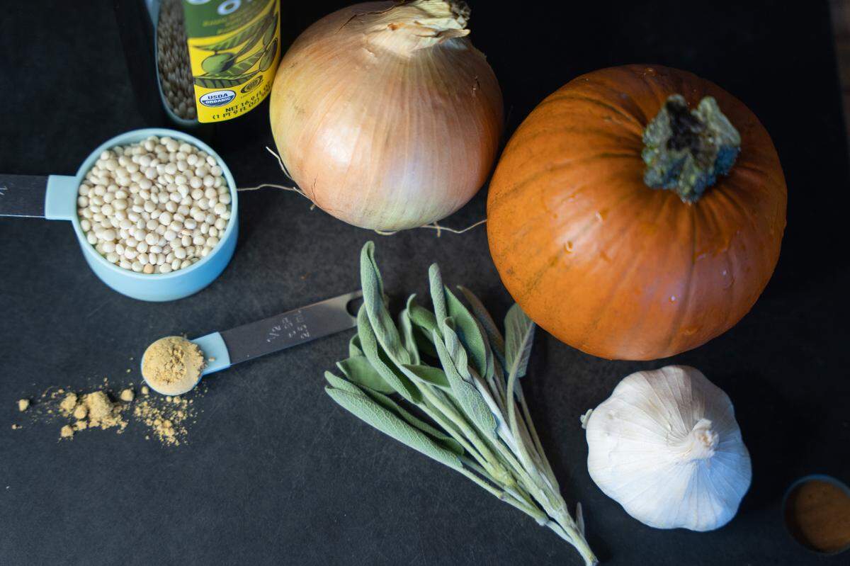 Ingredients for roast pumpkin soup on a cutting board, including a pumpkin, garlic, onion, sage, couscous and ground ginger.