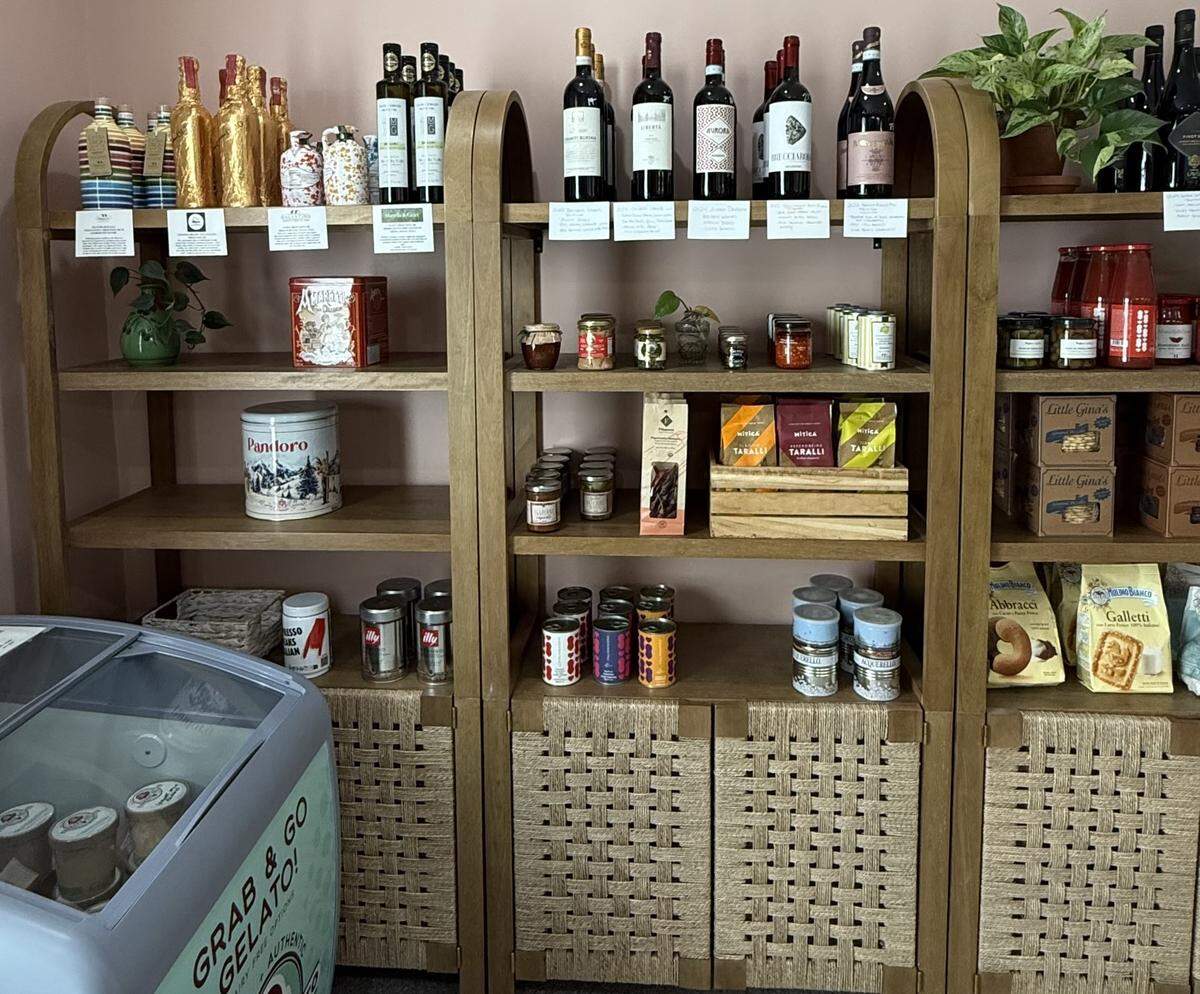 Wooden shelving units inside a shop filled with Italian specialty goods, including wine bottles, pasta sauces, canned tomatoes, and bags of “Little Gina’s” snacks.