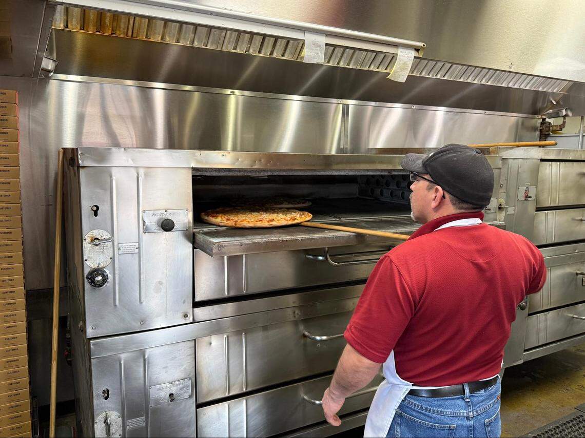 A cook in a red polo shirt and baseball cap uses a long wooden pizza peel to slide a fresh pizza into a large, industrial stainless steel deck oven in a professional kitchen setting.