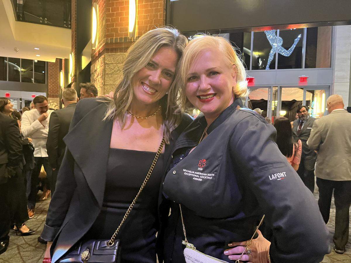 A close-up shot of two people smiling and posing together at an upscale indoor event. The one on the left has long blonde hair and is wearing a black blazer. The one on the right has short blonde hair and is wearing a black chef-style jacket embroidered with a “Michelin American South Regional Cocktail Awards” logo. The background is filled with other people mingling.