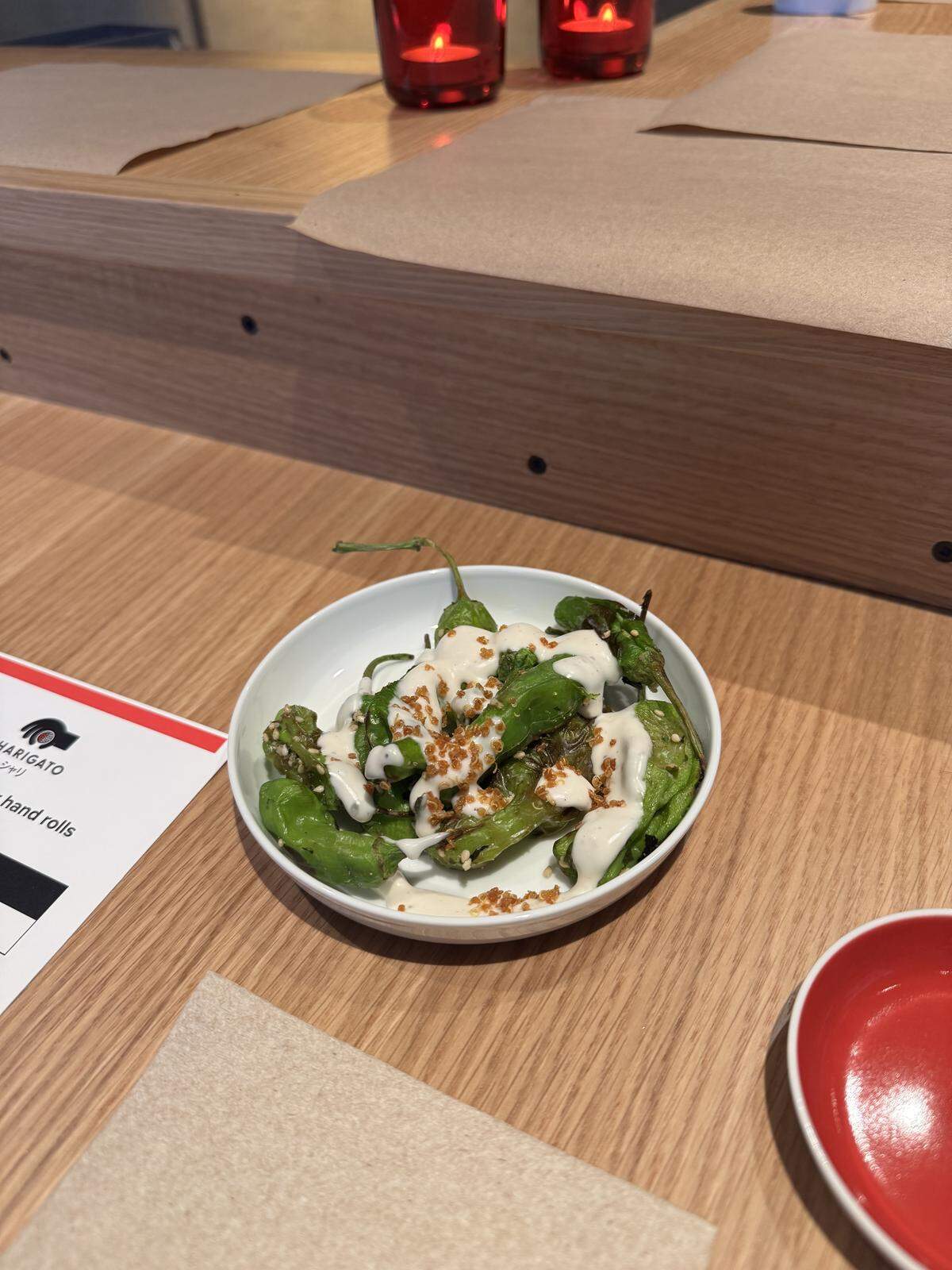 A small white bowl of charred green shishito peppers sits on a light wooden table. The peppers are drizzled with a creamy white sauce and topped with toasted sesame seeds and crunchy golden-brown bits. In the background, a menu with “hand rolls” text and two red votive candles are visible.