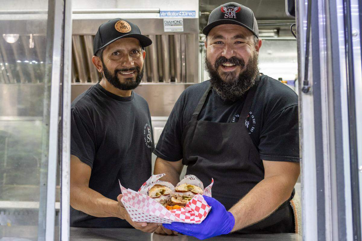 A portrait of two smiling chefs inside their food truck, the 5th Sin Wood Fire Kitchen. The man in front, with a full beard, wears a blue glove and holds a basket of sliders. The man behind him smiles as well. They are framed by the service window of their professional, stainless steel mobile kitchen.