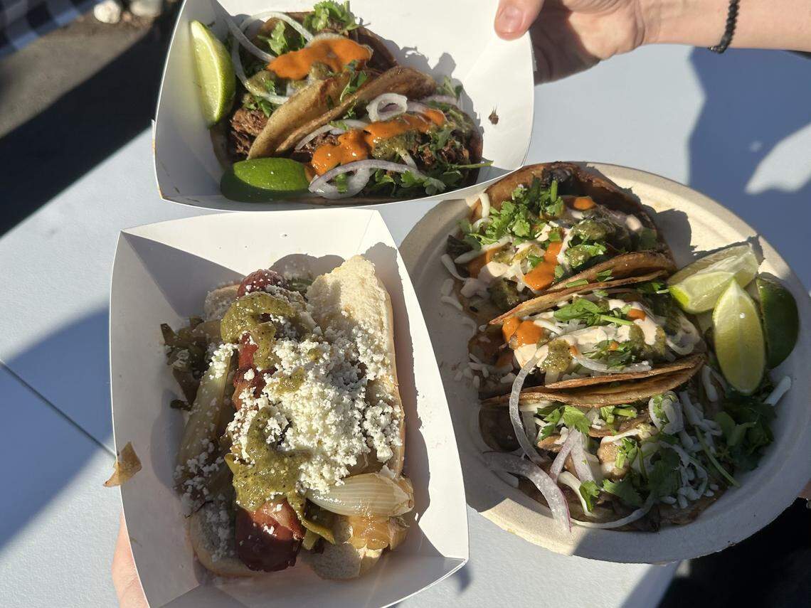 A person’s hands are holding three dishes of food outdoors on a white table. The dishes include: a paper tray of two tacos topped with meat, white onion, cilantro, and orange sauce, with a lime wedge; a paper tray containing a hot dog in a bun topped with grilled onions, green sauce, and crumbled white cheese; and a round plate holding three tacos topped with meat, white onion, green sauce, white sauce, and cilantro, served with two lime wedges.