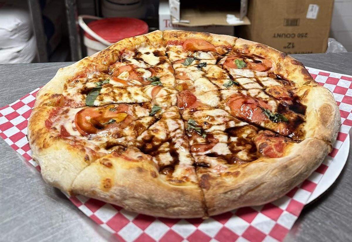 A freshly baked pizza, sliced into eight pieces, is presented on a stainless steel counter. It is a Caprese-style pizza with a hand-tossed crust, tomato sauce, mozzarella, sliced fresh tomatoes, and basil, all drizzled generously with a thick balsamic glaze.