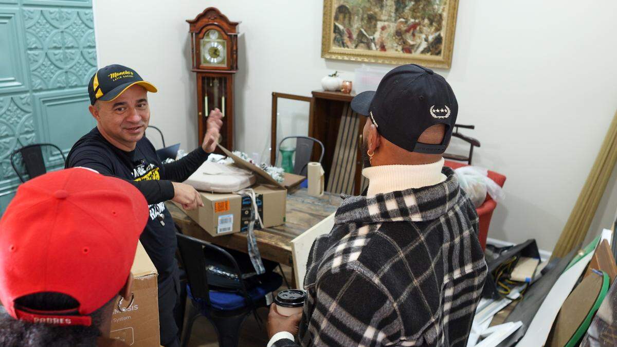 A person in a black t-shirt and cap speaks to two individuals in a room containing a grandfather clock, a wooden table with cardboard boxes, and a framed painting. One person in the foreground is wearing a black and white plaid jacket and holding a coffee cup.