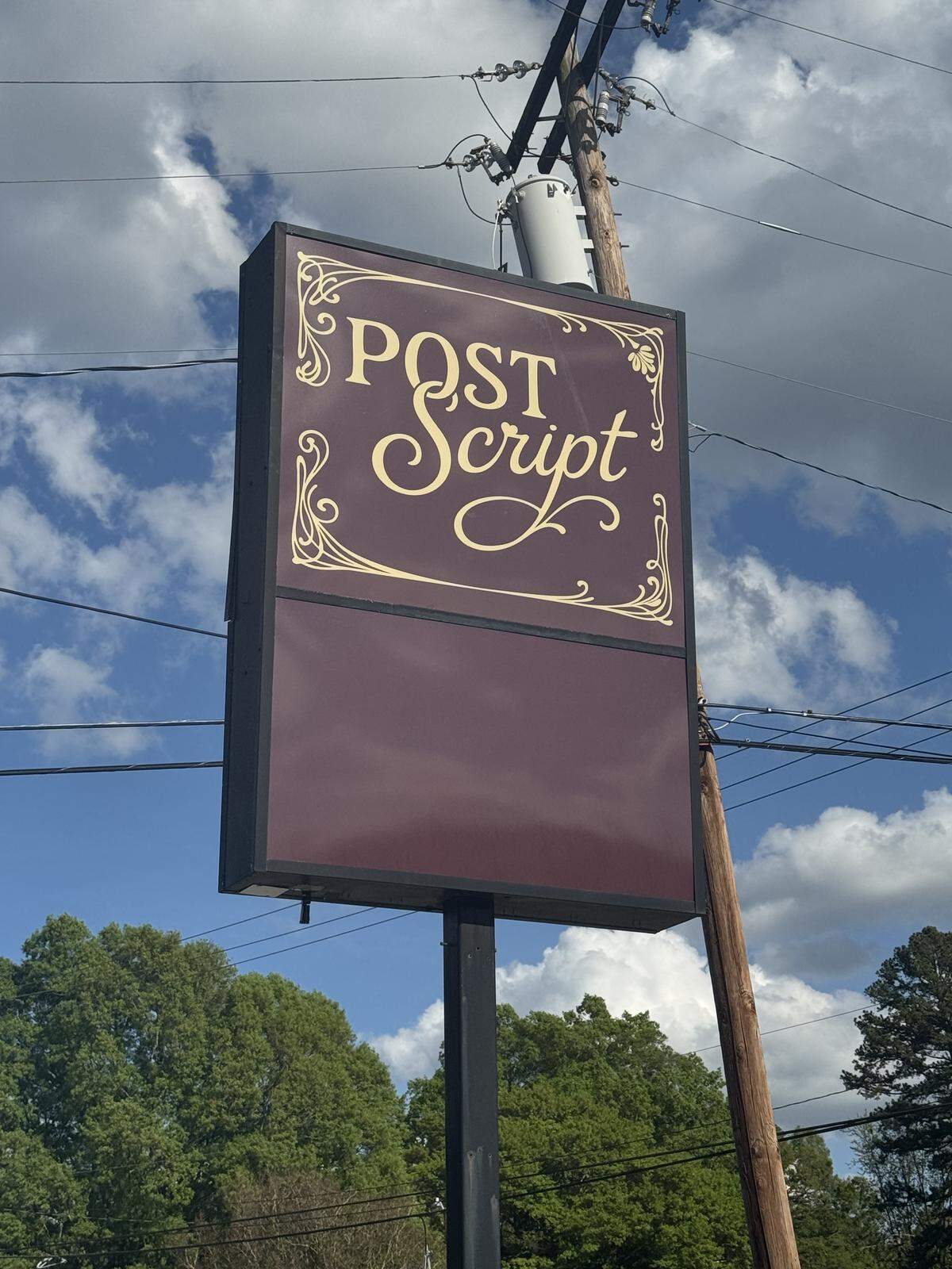 A tall outdoor sign against a blue sky with white clouds. The sign is dark burgundy with a gold ornate border and reads “Post Script” in an elegant, gold script font.